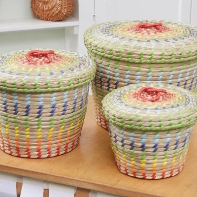 Três cestos redondos coloridos de palha com tampas e alças vermelhas sobre uma mesa de madeira