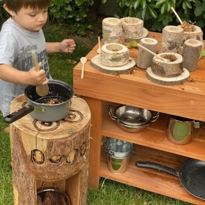Criança a brincar com cozinha de brinquedo de madeira e utensílios de metal num jardim.
