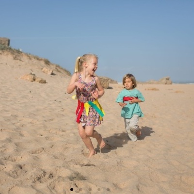 Duas crianças brincando na praia, menina com vestido colorido e menino com camisa azul.