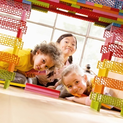 Três crianças deitadas olhando através de uma estrutura colorida feita de blocos transparentes.