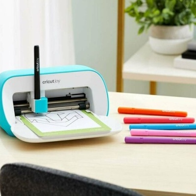 Máquina de corte Cricut Joy branca e azul claro com canetas coloridas numa mesa