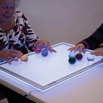 Mesa branca iluminada com moldura LED e bolas coloridas nas mãos de duas pessoas
