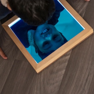 Espelho quadrado com moldura de madeira refletindo rosto de criança no chão.
