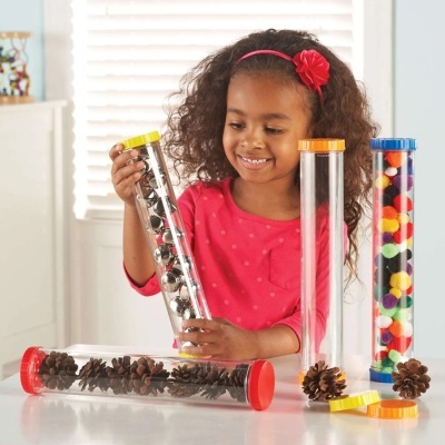 Menina segurando tubo transparente com bolas prateadas, outros tubos na mesa com pinhas e bolas coloridas