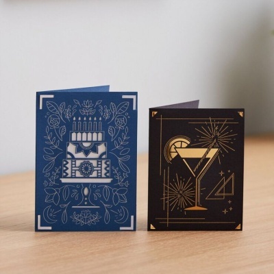 Dois cartões dobráveis decorativos, um azul com bolo de aniversário e outro preto com cocktail e fogos-de-artifício