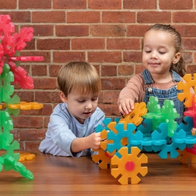 Crianças a brincar com blocos de construção de plástico coloridos numa mesa de madeira com parede de tijolos ao fundo.