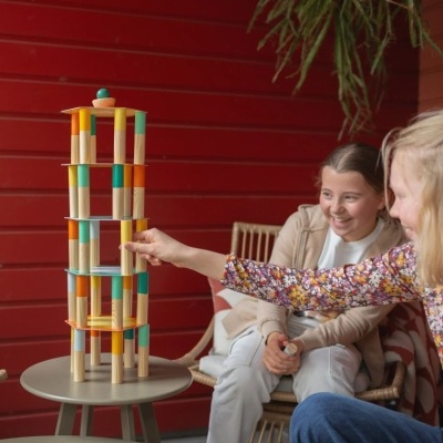 Torre de blocos de madeira coloridos em mesa com crianças ao lado e parede vermelha ao fundo