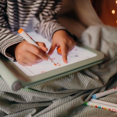 Criança a desenhar num tablet com capa verde claro, sentada com pijama riscado cinza e branco