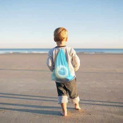 Criança de costas na praia com mochila saco azul transparente e brinquedo azul interno