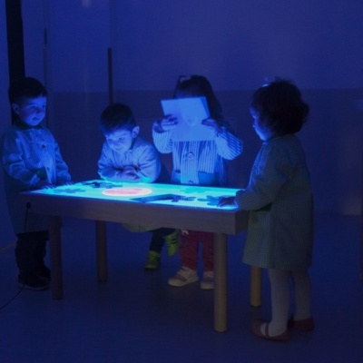 Crianças a brincar numa mesa iluminada de branco e azul num ambiente escuro