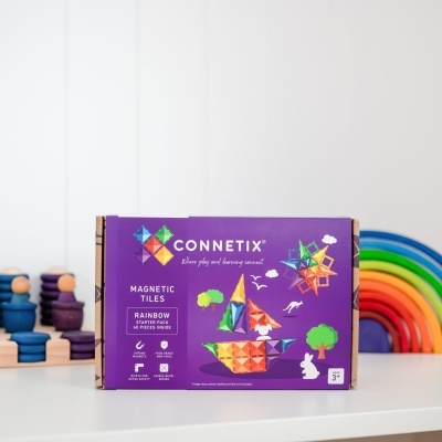 Caixa de azulejos magnéticos Connetix Rainbow Starter Pack sobre uma prateleira branca com brinquedos coloridos ao fundo