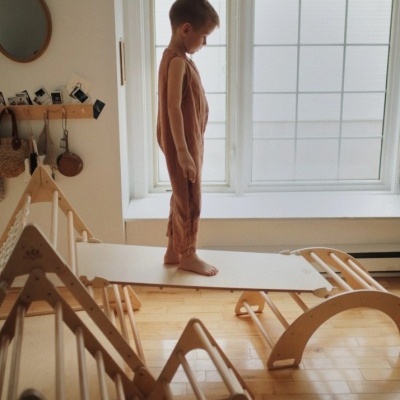 Criança em roupa castanha cor-de-rosa em prancha branca sobre estrutura de madeira clara num quarto com madeira clara e janela grande