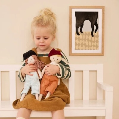 Criança segurando duas bonecas de tecido sentada num banco branco com parede bege e quadro decorativo ao fundo