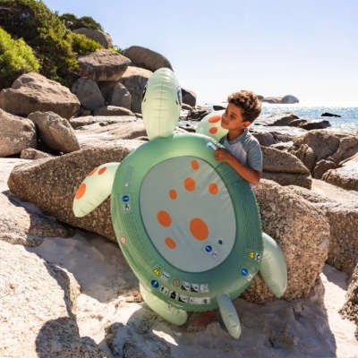 Bóia insuflável verde em forma de tartaruga com manchas laranja na praia com pedras grandes e criança