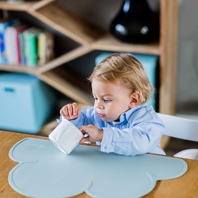 Criança pequena com copo branco numa mesa de madeira com toalha azul em forma de nuvem