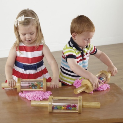 Crianças a usar rolos de madeira com cilindros transparentes e peças coloridas sobre massa rosa numa mesa de madeira