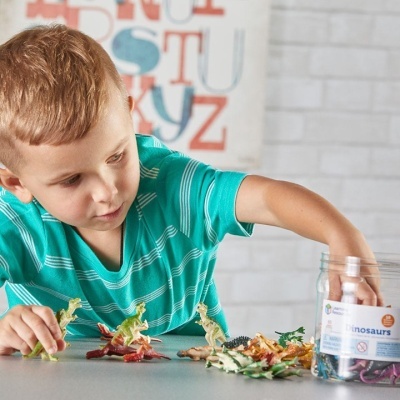 Menino brinca com dinossauros de brinquedo coloridos sobre mesa com frasco de dinossauros ao lado