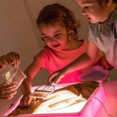 Crianças a brincar com areia numa caixa de luz retangular rosa