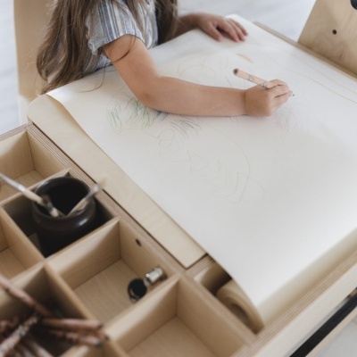 Criança a desenhar numa secretária de madeira clara com compartimentos organizadores.