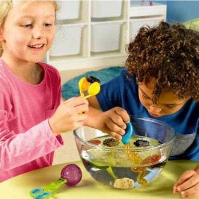 Crianças a brincar com recipiente de vidro cheio de água e acessórios coloridos numa mesa amarela