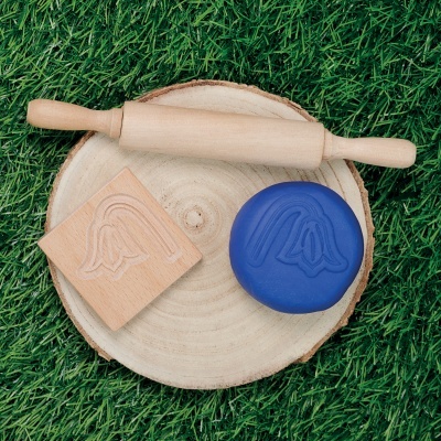 Conjunto de ferramentas para modelar massa com rolo, carimbo de madeira e massa azul com padrão em relevo, sobre disco de madeira em relva verde.