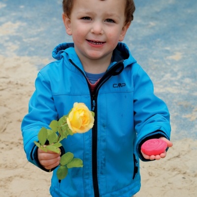 Criança com casaco azul segurando flor amarela e brinquedo rosa em parque com areia