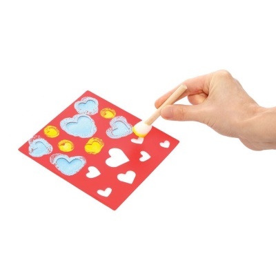 Mão aplica tinta amarela num molde vermelho com corações recortados.