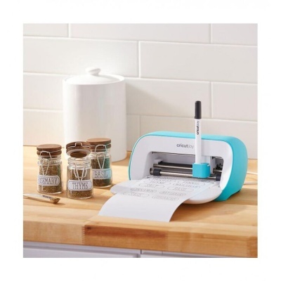 Aparelho Cricut Joy branco e azul numa bancada de madeira ao lado de frascos rotulados com especiarias numa cozinha