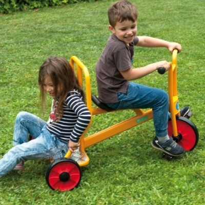 Duas crianças a brincar num triciclo amarelo com rodas vermelhas num campo relvado