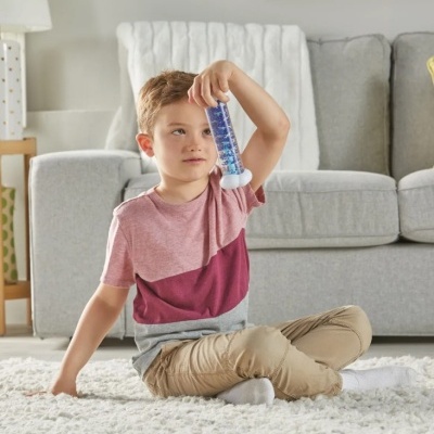 Menino segurando cilindro de líquido azul sentado em tapete branco com sofá ao fundo