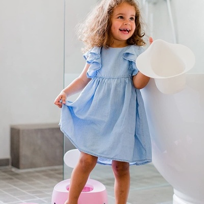 Menina com vestido azul claro e urinol infantil branco na casa de banho, penico rosa no chão
