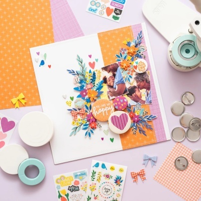 Álbum de fotos decorado com flores de papel, corações e adesivos coloridos em fundo branco, laranja e roxo, com materiais de scrapbook ao redor.