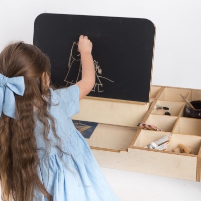Menina a desenhar num quadro negro de uma mesa em madeira clara com divisórias e materiais de desenho.