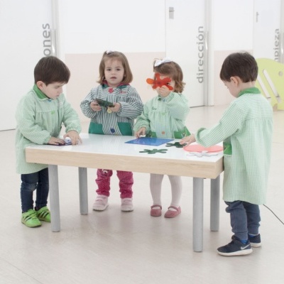 Crianças vestindo aventais verdes à volta de uma mesa em sala branca com portas ao fundo.