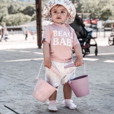Criança com chapéu floral e t-shirt rosa 'BEACH BABY' segurando baldes rosa na areia