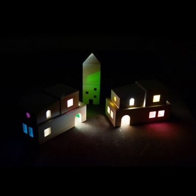 Miniaturas de casas e torre iluminadas interiormente em várias cores, num fundo escuro.