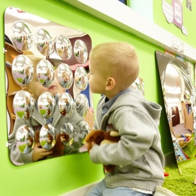 Espelho convexo com 20 semiesferas refletindo várias imagens, parede verde, criança ajoelhada com brinquedo.