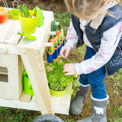 Carrinho de madeira para jardinagem infantil com plantas e acessórios, criança a brincar.