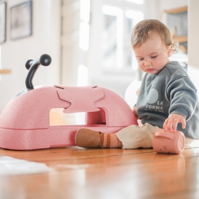 Bebé sentado no chão ao lado de trenó cor de rosa com guiador preto.