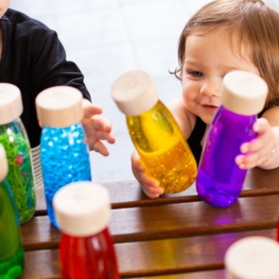 Crianças brincando com garrafas sensoriais coloridas de plástico numa mesa de madeira