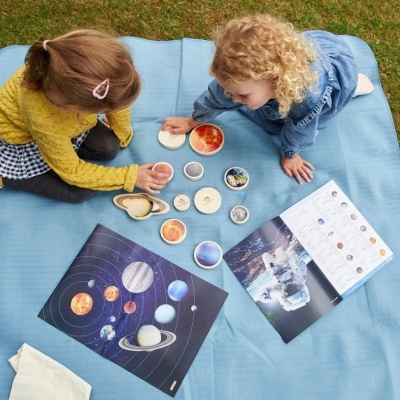 Crianças brincando com cartas dos planetas do sistema solar e astronautas num cobertor azul no relvado