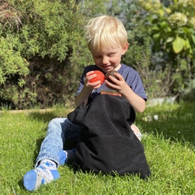 Criança loira sentada na relva a segurar duas bolas coloridas com mochila preta no colo
