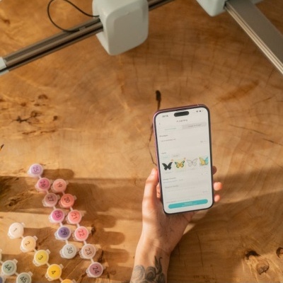 Mão segurando smartphone com imagens de borboletas na tela e frascos de tinta numerados sobre mesa de madeira.