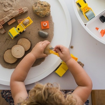Criança a brincar com brinquedos de construção amarelos, mesa branca com compartimentos e areia