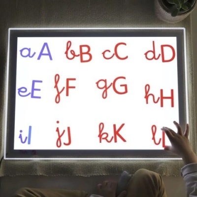 Quadro luminoso com letras do alfabeto vermelho e azul, criança a tocar e planta ao lado.