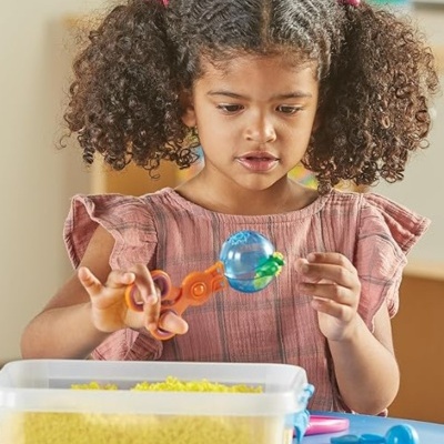Menina a brincar com pinça e bola azul sobre recipiente com arroz amarelo