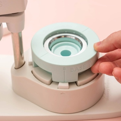 Base giratória cinza claro com anel metalizado e mão ajustando, apoio branco e fundo rosa claro