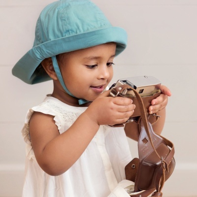 Criança com chapéu azul e vestido branco segurando câmara fotográfica vintage de pele castanha.