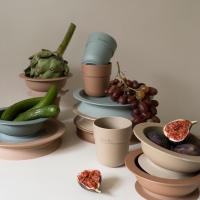 Conjunto de louça em tons pastel com frutas e legumes, sobre superfície branca e fundo bege