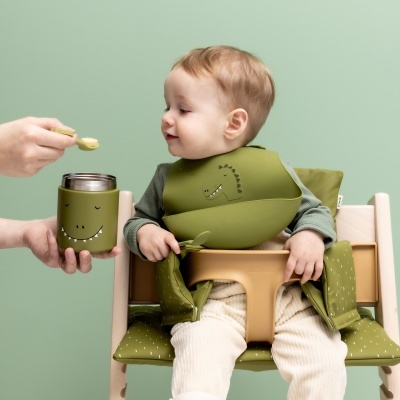 Bebé com babete verde a ser alimentado com colher e recipiente térmico com rosto sorridente.